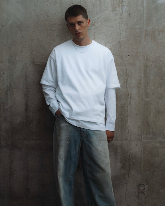 Person wearing a white layered top and light-wash jeans from the solebox Core Collection, standing against a concrete wall.