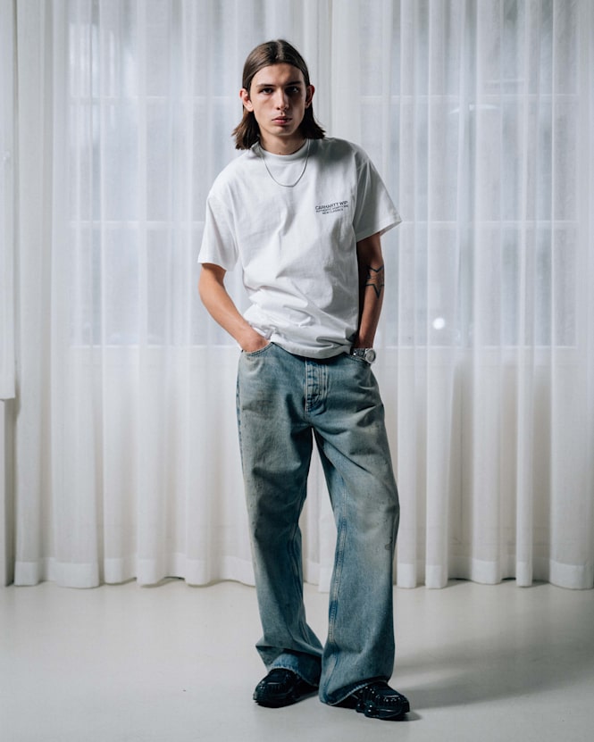 Full-body view of a person wearing a white Carhartt WIP T-shirt with small chest logo, loose light blue jeans, and black shoes, standing with hands in pockets in front of sheer white curtains.