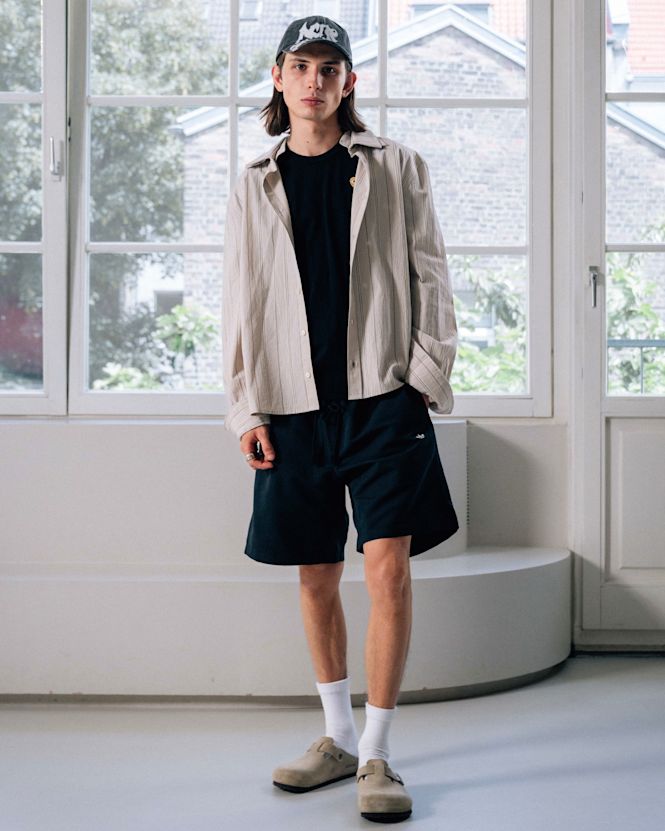 Mid-length view of a person wearing a light striped overshirt over a black T-shirt, paired with black shorts, white socks, and beige clogs, styled with a cap, standing in front of a window backdrop.