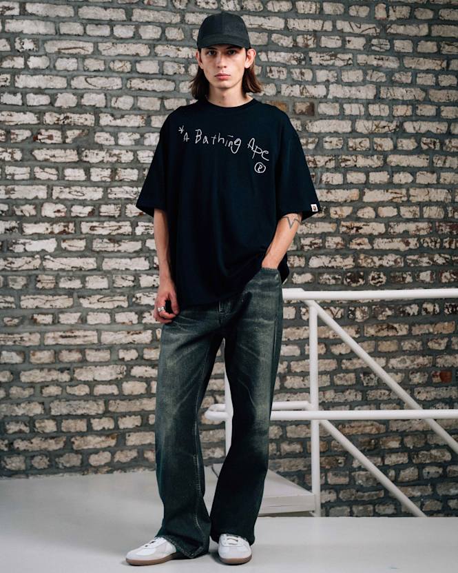 Full-body view of a person wearing a black A Bathing Ape T-shirt with handwritten-style logo, dark denim jeans, white sneakers, and a black cap, standing in front of a brick wall.