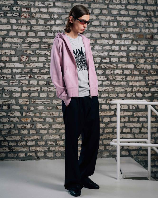 Mid-length view of a person wearing a pink zip-up hoodie over a graphic T-shirt, styled with black wide-leg pants and tinted sunglasses, standing in front of a brick wall background.