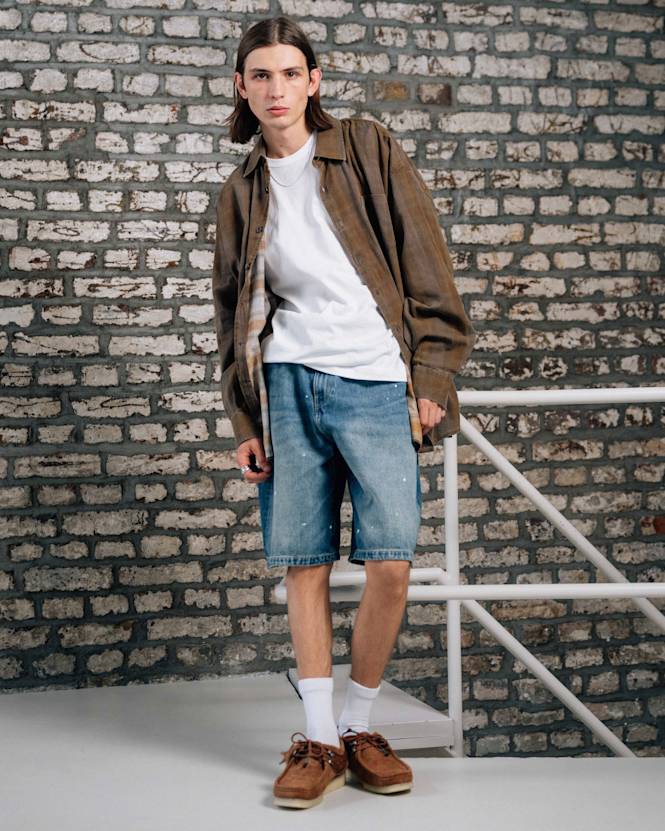 Full-body view of a person wearing a brown overshirt layered over a white T-shirt, paired with light blue denim shorts, white socks, and brown shoes, standing against a textured brick wall with a stair railing.