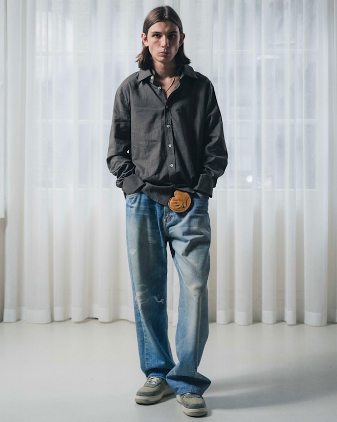 Full-body view of a person wearing a dark button-up shirt with light blue loose-fit jeans and sneakers, styled with a belt pouch detail, standing in front of sheer white curtains.