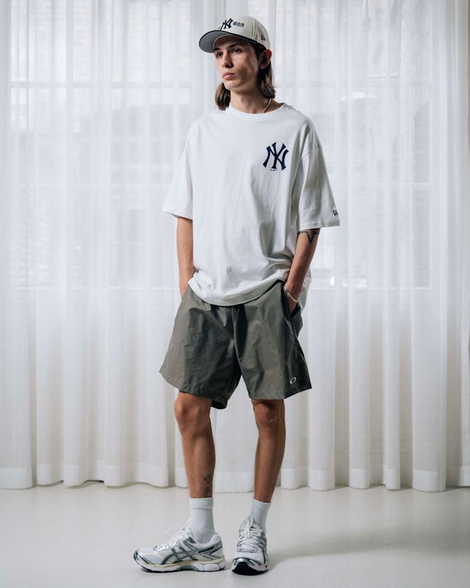 Mid-length view of a person wearing a white New York Yankees T-shirt with logo, olive green shorts, and a light-colored NY cap, styled with running sneakers against sheer curtain backdrop.
