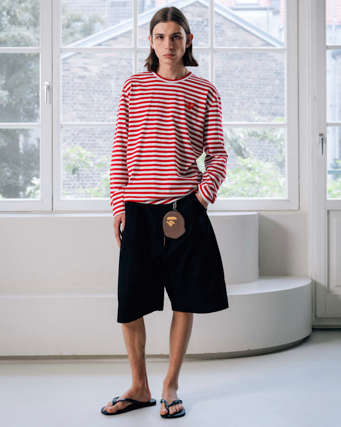 Full-body view of a person wearing a red-and-white striped long-sleeve shirt with a small heart patch, black shorts, and black flip-flops, standing indoors in front of a large window.