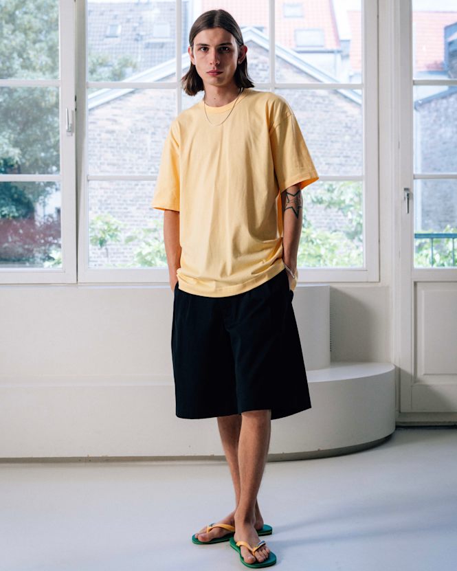 Full-body view of a person wearing a yellow T-shirt, black shorts, and green flip-flops, standing indoors in front of large windows with natural light.