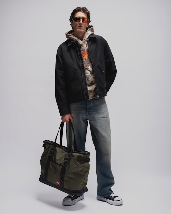 Person wearing a black workwear jacket over a camouflage hoodie, faded blue jeans, black-and-white sneakers, and mirrored sunglasses, carrying a large olive-green duffel bag against a light grey studio background.