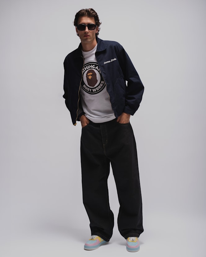 Person wearing a navy Human Made jacket over a white graphic T-shirt, loose dark denim jeans, pastel-colored sneakers, and black sunglasses against a light grey studio background.