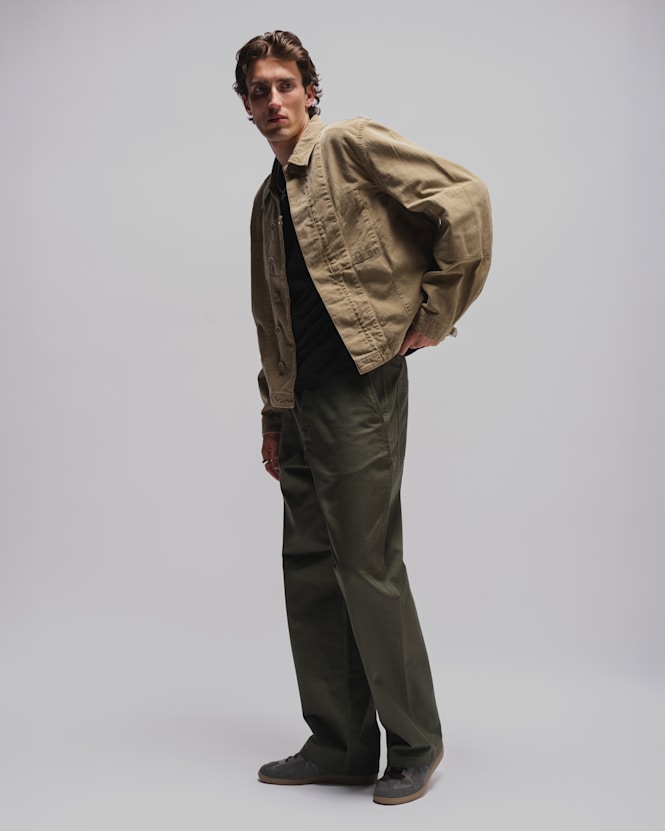 Person wearing a beige workwear jacket over a black T-shirt, olive green utility trousers, and grey suede shoes against a light grey studio background.  Geben Sie Ihr Feedback zu BizChat ab.