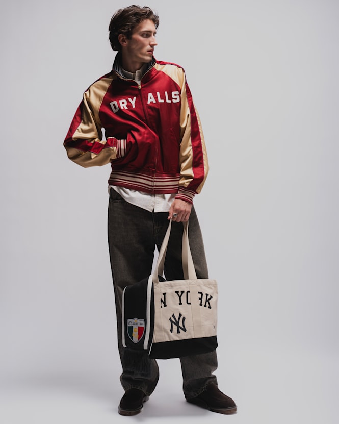 Person wearing a red and gold satin varsity jacket, faded black jeans, dark shoes, and carrying a New York Yankees tote bag against a light grey studio background.  Geben Sie Ihr Feedback zu BizChat ab.