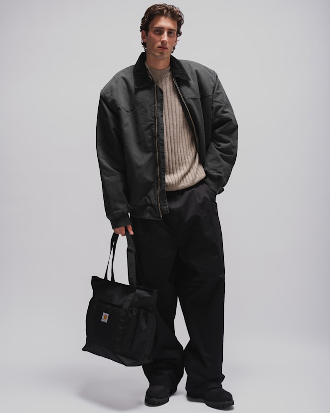 Person wearing a black workwear jacket, beige rib-knit sweater, loose black trousers, black boots, and carrying a black Carhartt tote bag against a light grey studio background.