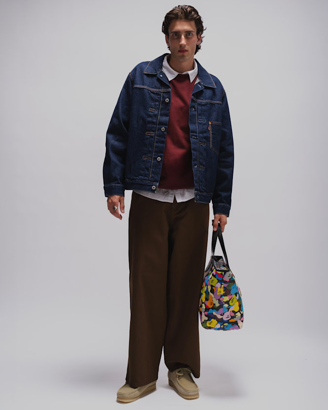 Person wearing a dark blue denim jacket over a burgundy sweater and white shirt, paired with wide-leg brown trousers, beige suede shoes, and carrying a colorful patterned tote bag against a light grey studio background.