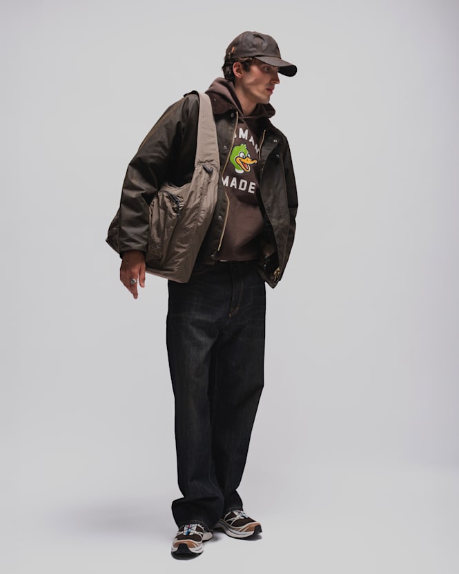 Person wearing a dark brown jacket over a graphic hoodie, dark blue jeans, a brown baseball cap, and running sneakers, carrying a large shoulder bag against a light grey studio background.