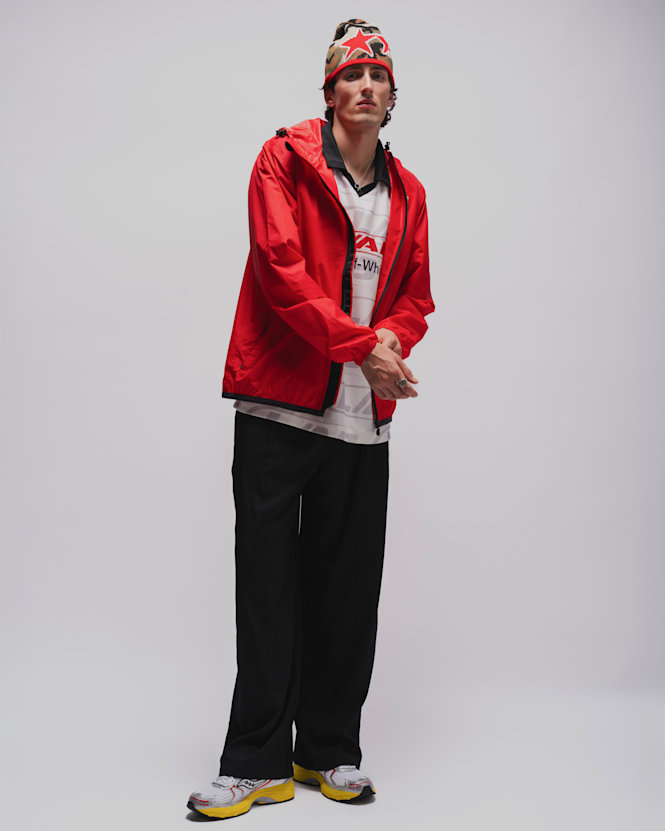 Person wearing a bright red lightweight jacket over a graphic football-style jersey, loose black trousers, white and yellow running sneakers, and a patterned knit beanie against a light grey studio background.