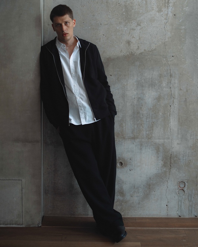 Person wearing a black zip-up jacket, white shirt, and black trousers from the solebox Core Collection, leaning against a concrete wall.