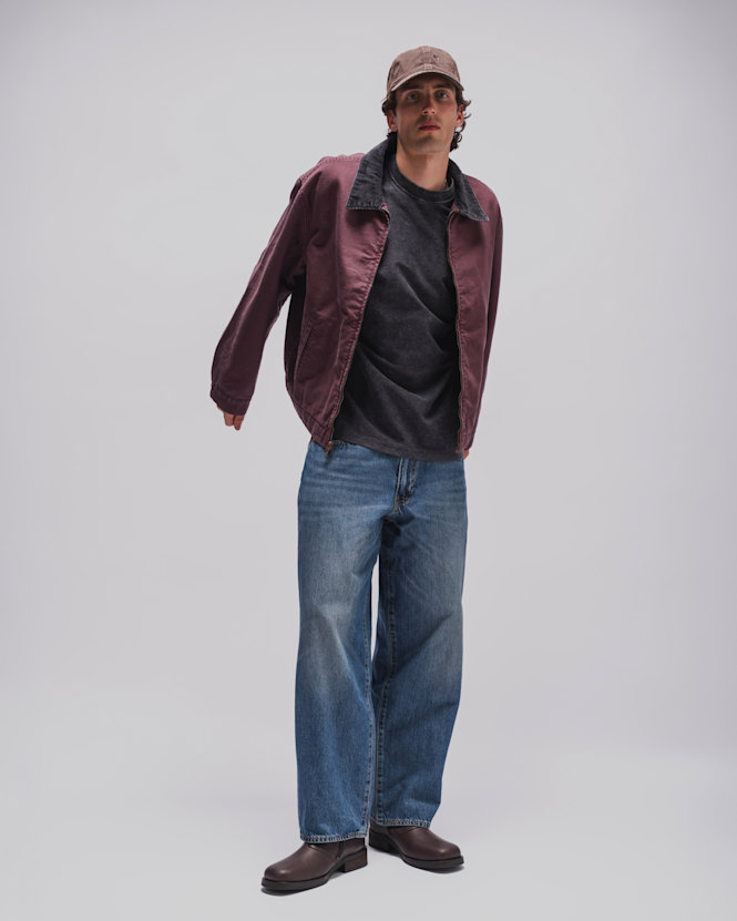 Person wearing a burgundy jacket over a faded dark grey T-shirt, light blue wide-leg jeans, brown leather boots, and a beige baseball cap against a light grey studio background.  Geben Sie Ihr Feedback zu BizChat ab.
