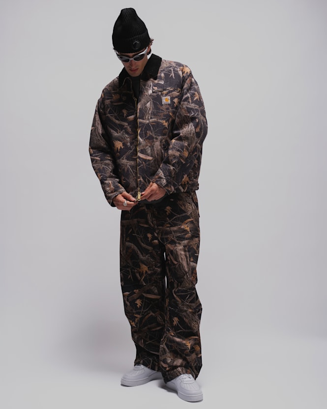 Person wearing a camouflage Carhartt workwear jacket and matching trousers, a black beanie, wraparound sunglasses, and white sneakers against a light grey studio background.