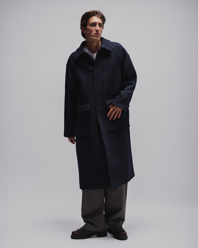 Person wearing a long navy coat over a white T-shirt, loose grey trousers, and dark leather shoes against a light grey studio background.